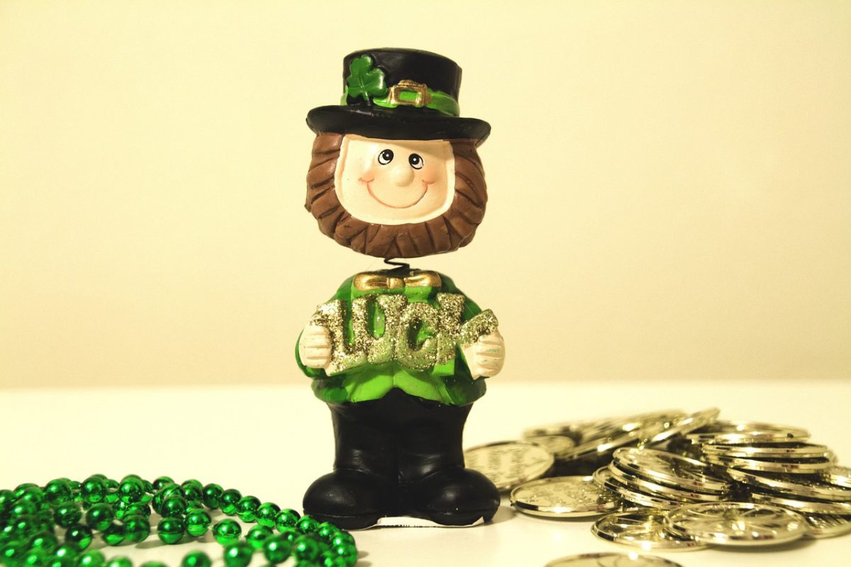 A leprechaun bobble head holding a sign that says, Luck is on a table surrounded by gold coins and a green necklace