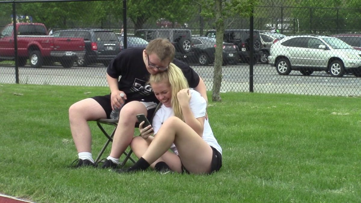 A high school boy sitting in a chair outside, hugging a high school girl on the ground next to him