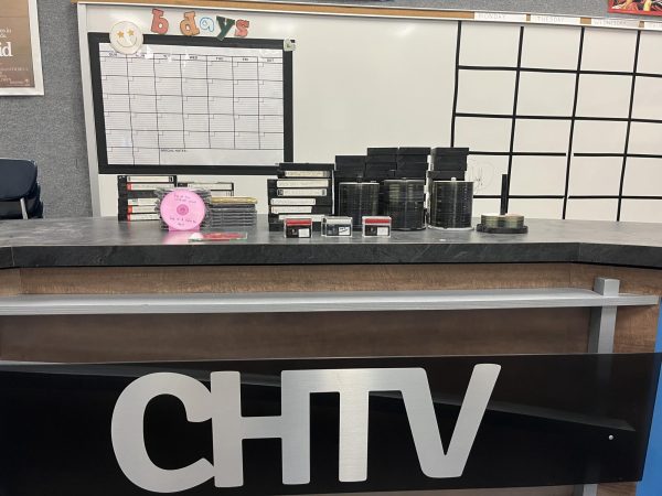 The CHTV news desk in front of a whiteboard in the classroom. On the desk is a lot of DVDs and VHSs.